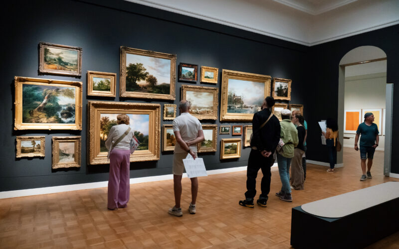 Een vijftal bezoekers kijkt naar een muur waarop een aantal geschilderde landschappen in grote gouden lijsten worden tentoongesteld.